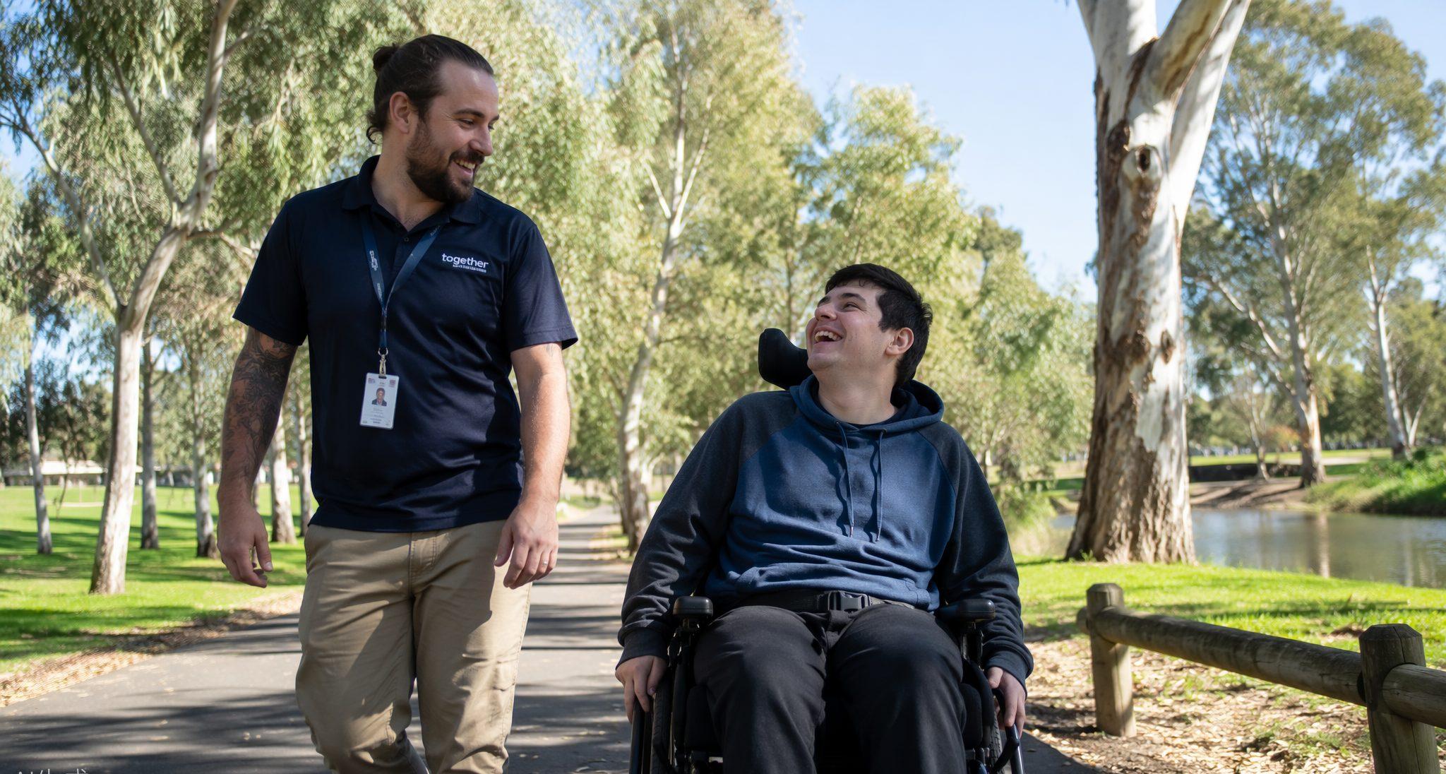 Support worker walking with a young man in a wheelchair through a sunny park
